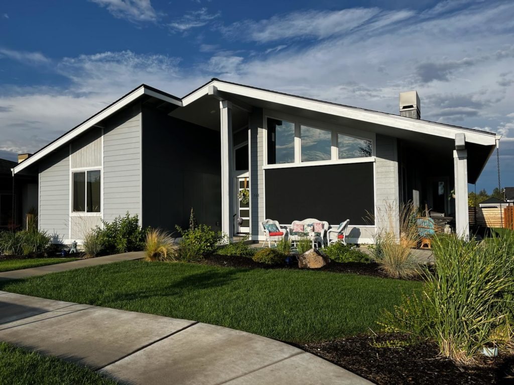 Black motorized patio screen lowered over a front porch seating area on a gray single-story home
